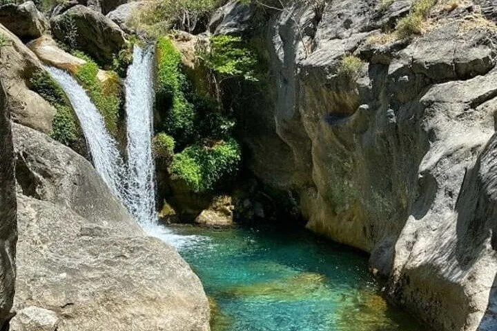 Ein kleiner Wasserfall fließt in ein klares Wasserbecken, umgeben von felsigem Gelände.