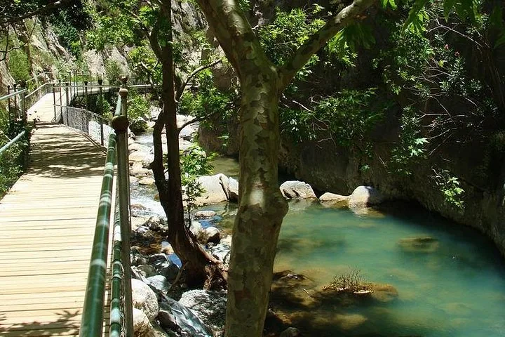 Ein Holzsteg, der am Rand eines türkisfarbenen Flusses durch eine schattige, felsige Schlucht verläuft.